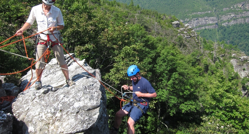 Two people wearing safety gear are secured by ropes near the edge of a cliff. One person appears to be an instructor, giving direction to the other person. There is a vast forested area below them. 